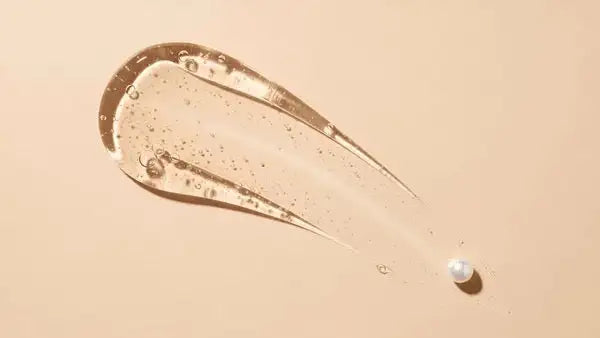 A clear, gel-like liquid with a subtle golden tint and tiny bubbles, spread in an elongated streak on a soft beige surface.